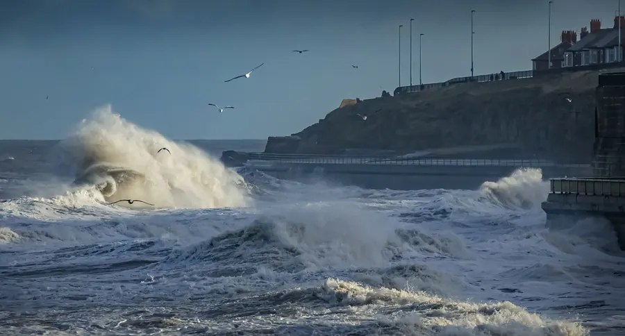 Storm waves breaking against a sea wall below seafront houses, the onshore conditions that lift loose lead flashings